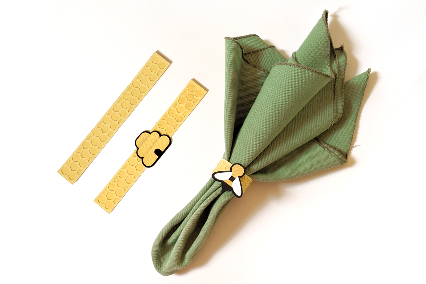 A sage green napkin on a white background. The napkin has a paper ring with a honeycomb pattern and a stylized simple bee. There are 2 more napkin rings laid flat nearby. One has a beehive, the other just has the honeycomb pattern.