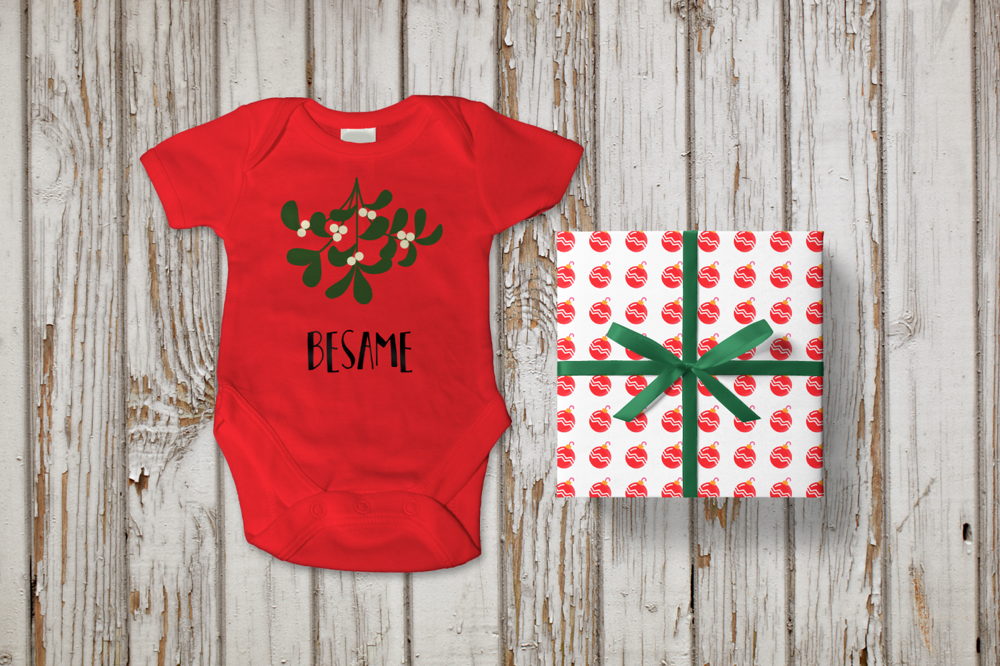 Red baby onesie on a weathered wood background with a wrapped gift next to it. The onesie has a sprig of mistletoe with the word "besame" below.