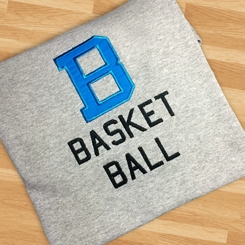 A folded grey heather shirt sits on a wood background. Embroidered onto the shirt is the word "basketball" in two lines. Above is a large applique B in blue fabric.