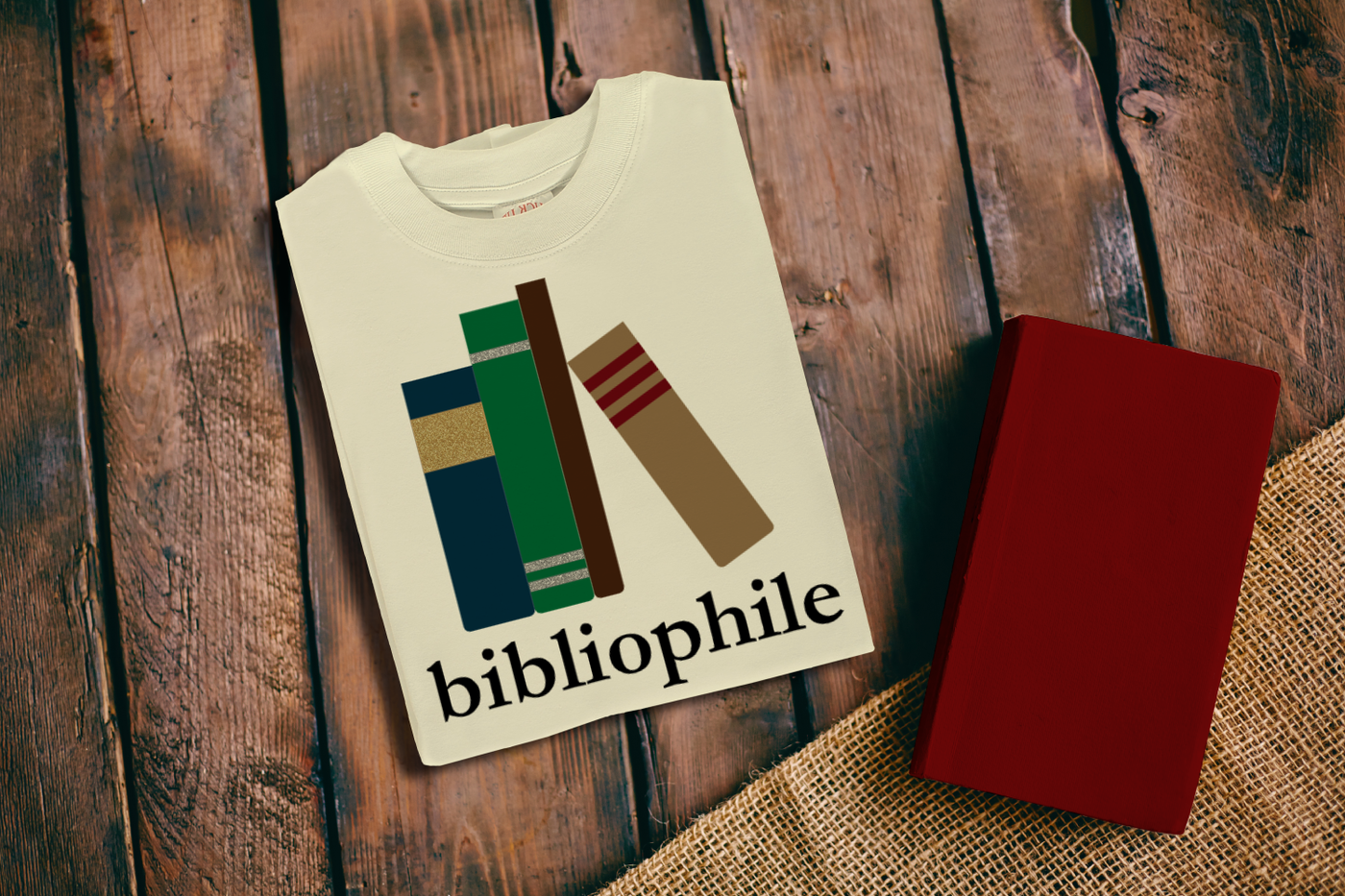A folded ivory shirt on a wood background with a red book laying nearby. The shirt has a design of 4 book spines and underneath is the word "bibliophile."