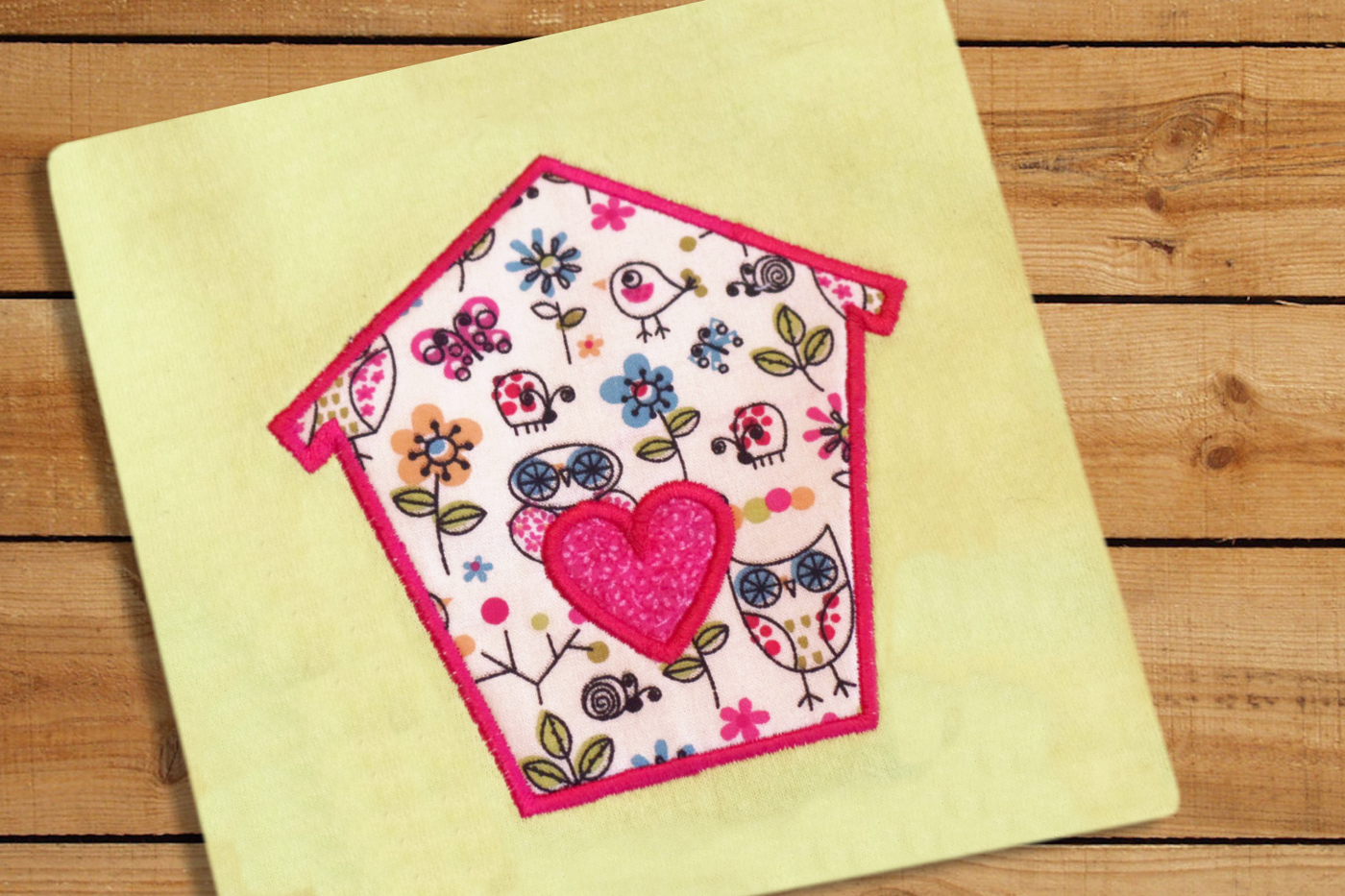 A yellow square of fabric on a wood surface. Embroidered onto the fabric is an applique birdhouse shape with a heart in the middle. The applique fabric features stylized birds.
