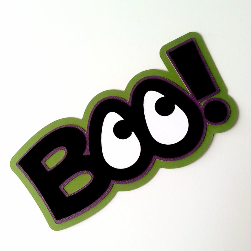 The word "BOO!" in black, white, green, and purple made out of layered paper. The Os look like eyes.