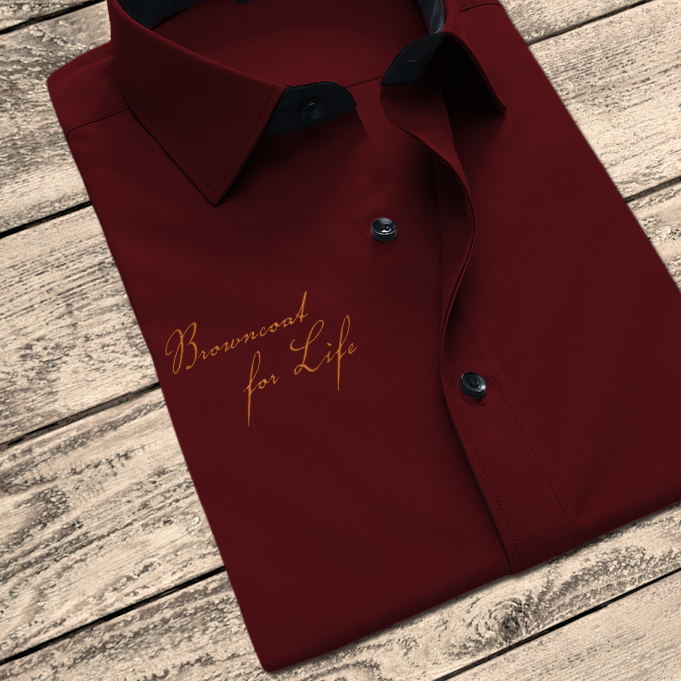 A maroon button up shirt on a wood background. Embroidered onto the shirt in orange is the phrase "Browncoat for Life" done in a scripty font.