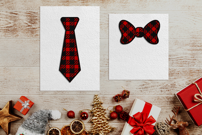 Two cards on a wood background with presents and Christmas decorations below. One card has a paper neck tie, the other has a paper bow tie. Each tie has a layered paper buffalo plaid pattern in red and black.