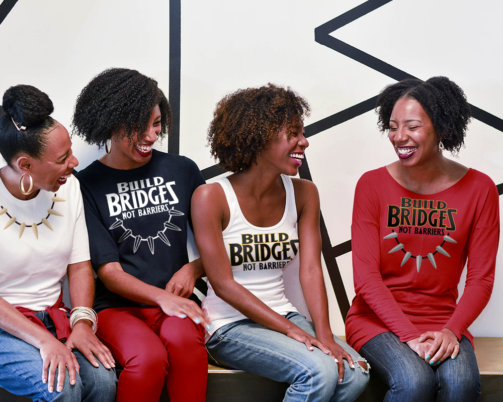 Four black women sit on a bench in front of a black and white wall. Each has a different design on their shirt. The woman on the left has a shirt with an animal tooth design. The other 3 women have the words "Build bridges not barriers." Two of these women also have the tooth necklace below the words.