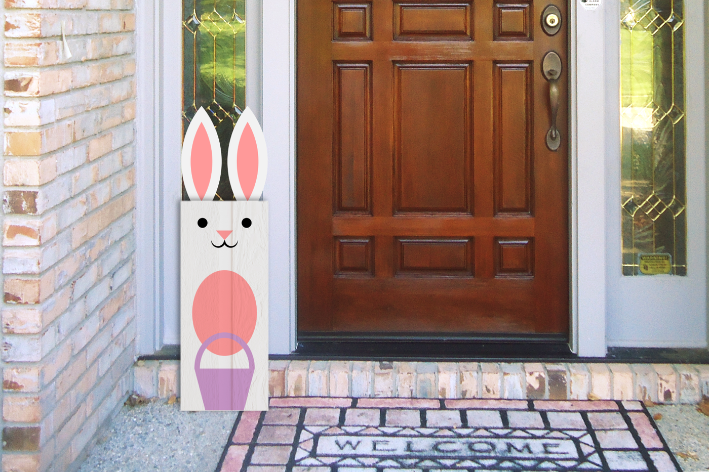 Front door of a house. In front is a white painted vertical wood sign decorated to look like an Easter bunny with ears at the top.