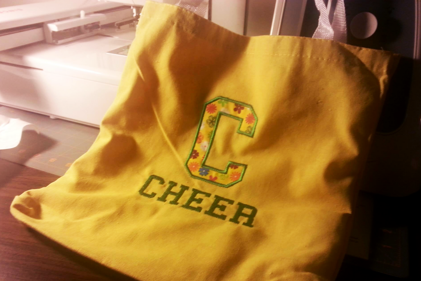 A yellow tote bag with the word "CHEER" embroidered onto it. Above is an applique letter C.