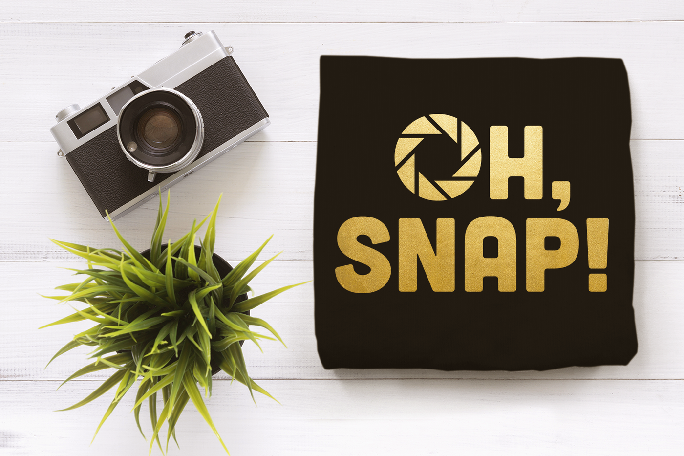"Oh Snap!" SVG design with an aperture in place of the O. Shown in gold on a black shirt. The shirt lays on a white wood surface with a camera and plant nearby.