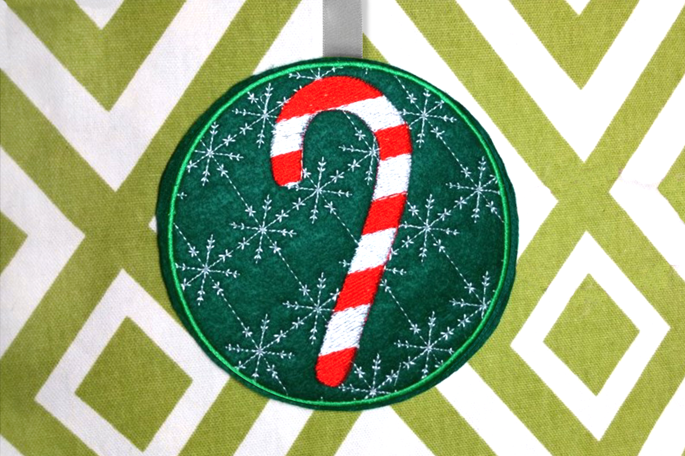 In the hoop Christmas ornament. A round green piece of felt has the embroidery design of a striped candy cane. In the background is an embroidered snowflake pattern, set inside of a circle. A ribbon has been added to the top.