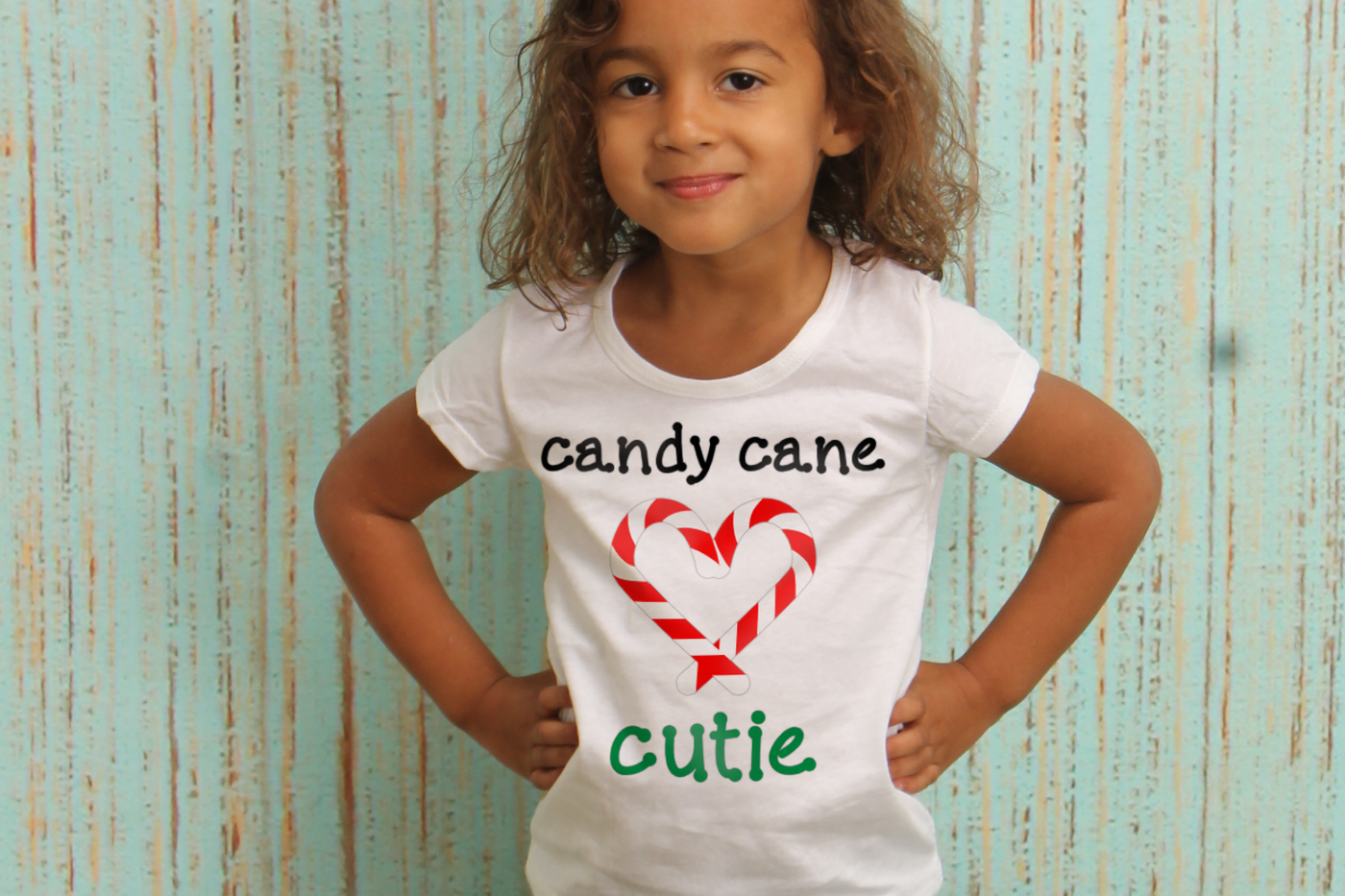 A black little girl smiles with her hands on her hips. Her shirt says "candy cane cutie" and there are 2 candy canes forming a heart shape.