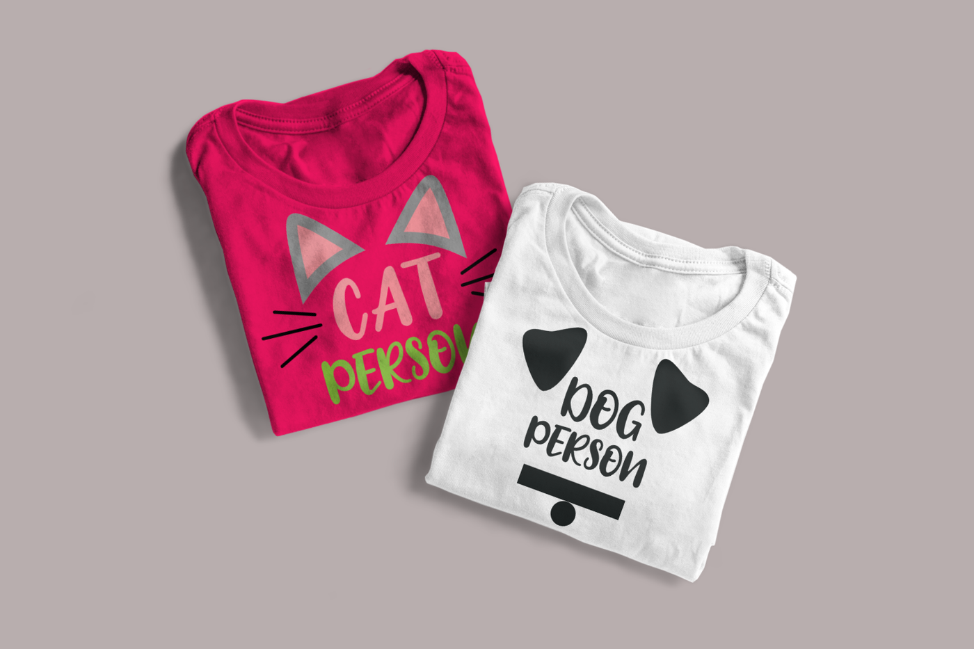 Two folded shirts on a grey background. One shirt says "Cat Person" with cat ears and whiskers, and the other says "Dog person" with dog ears and a collar with tag.