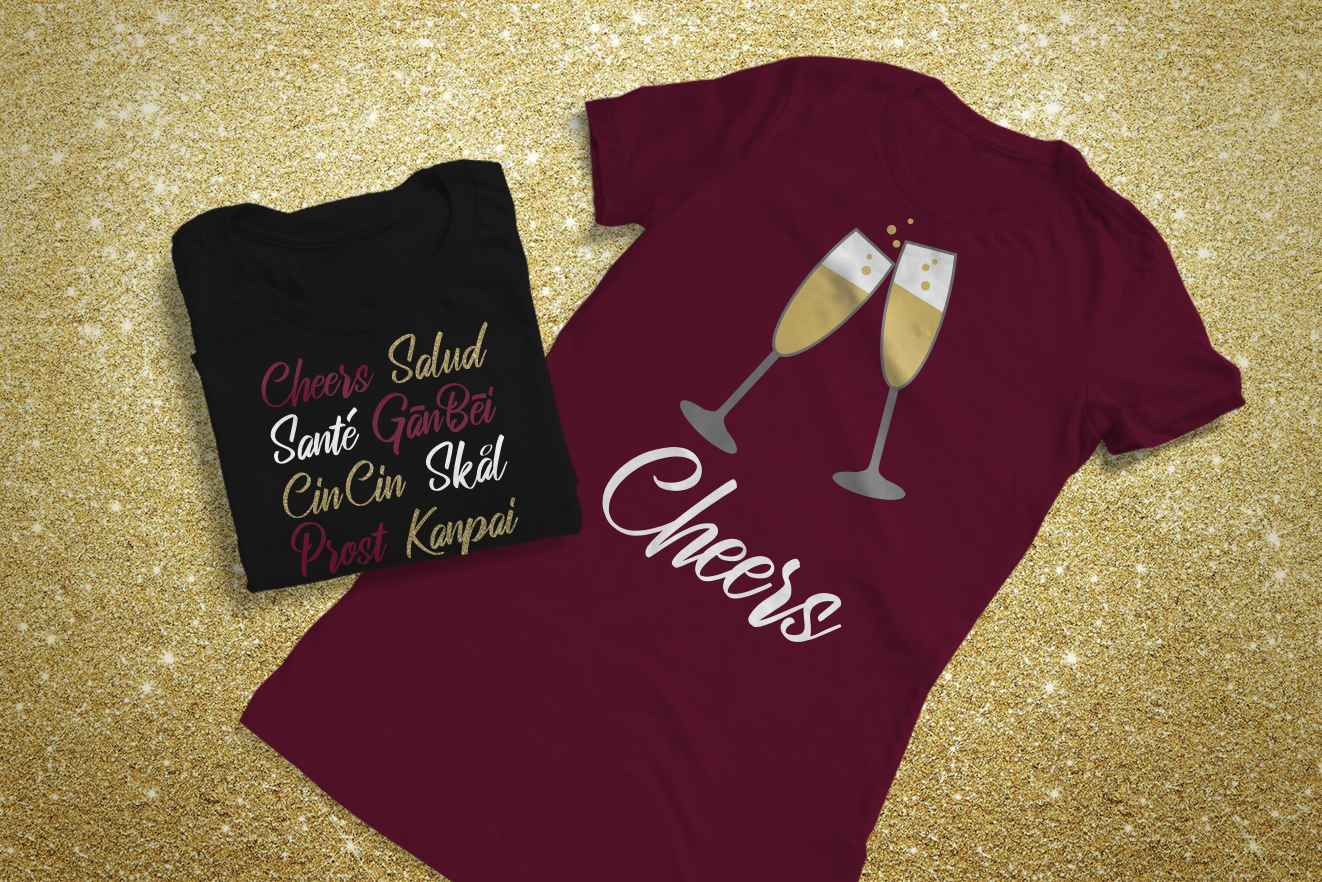 Two tees on a glittery gold background. The black tee is folded, the wine colored tee is not. The black tee has the words Cheers, Salud, Sante, Gan Bei, CinCin, Skal, Prost, and Kanpai in gold, white, and wine. The wine colored shirt has two toasting champagne flutes and the word "Cheers" in white.