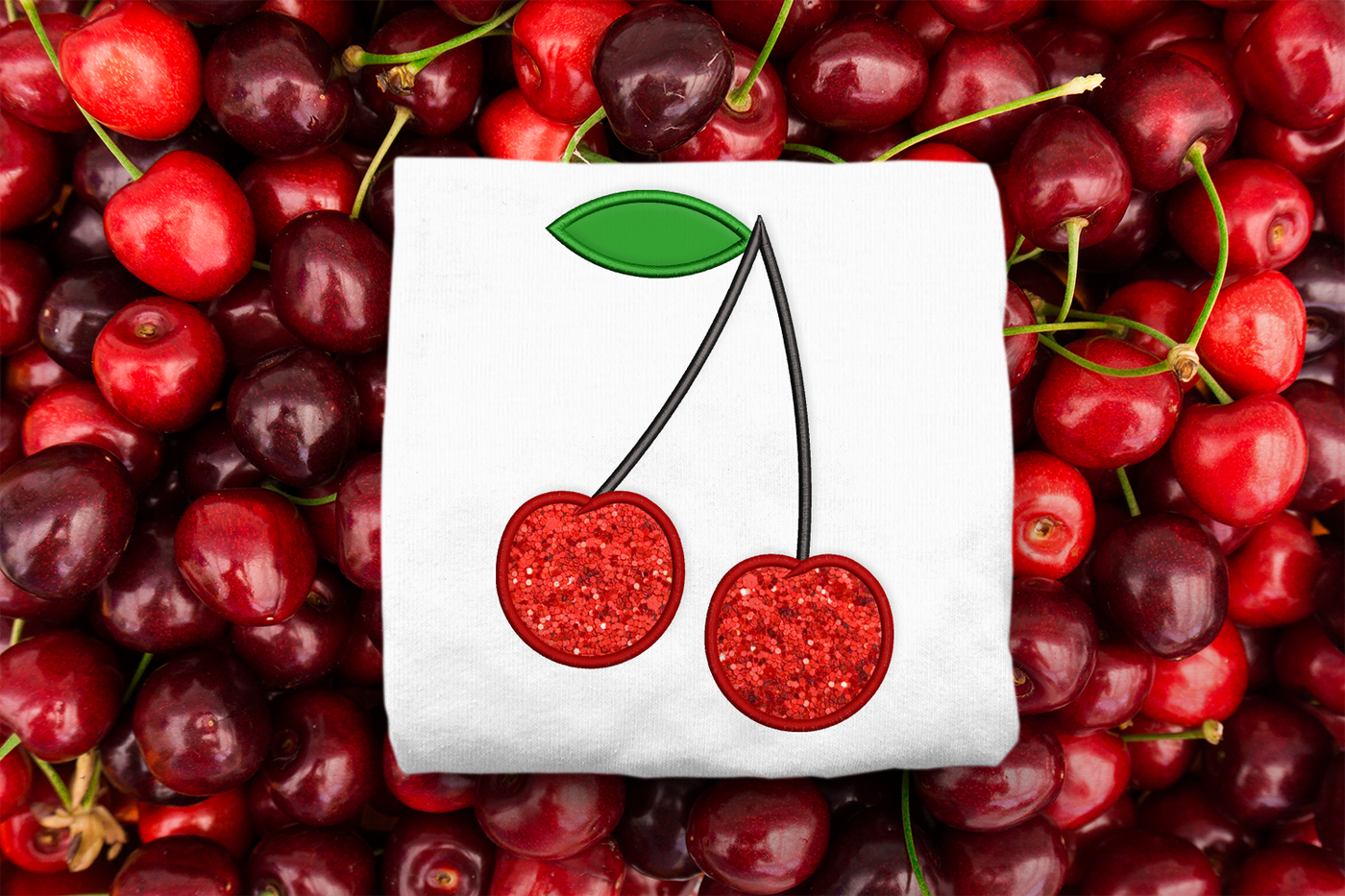 Folded white shirt on a background of cherries. On the shirt is an applique design of pair of cherries on the stem with a single leaf