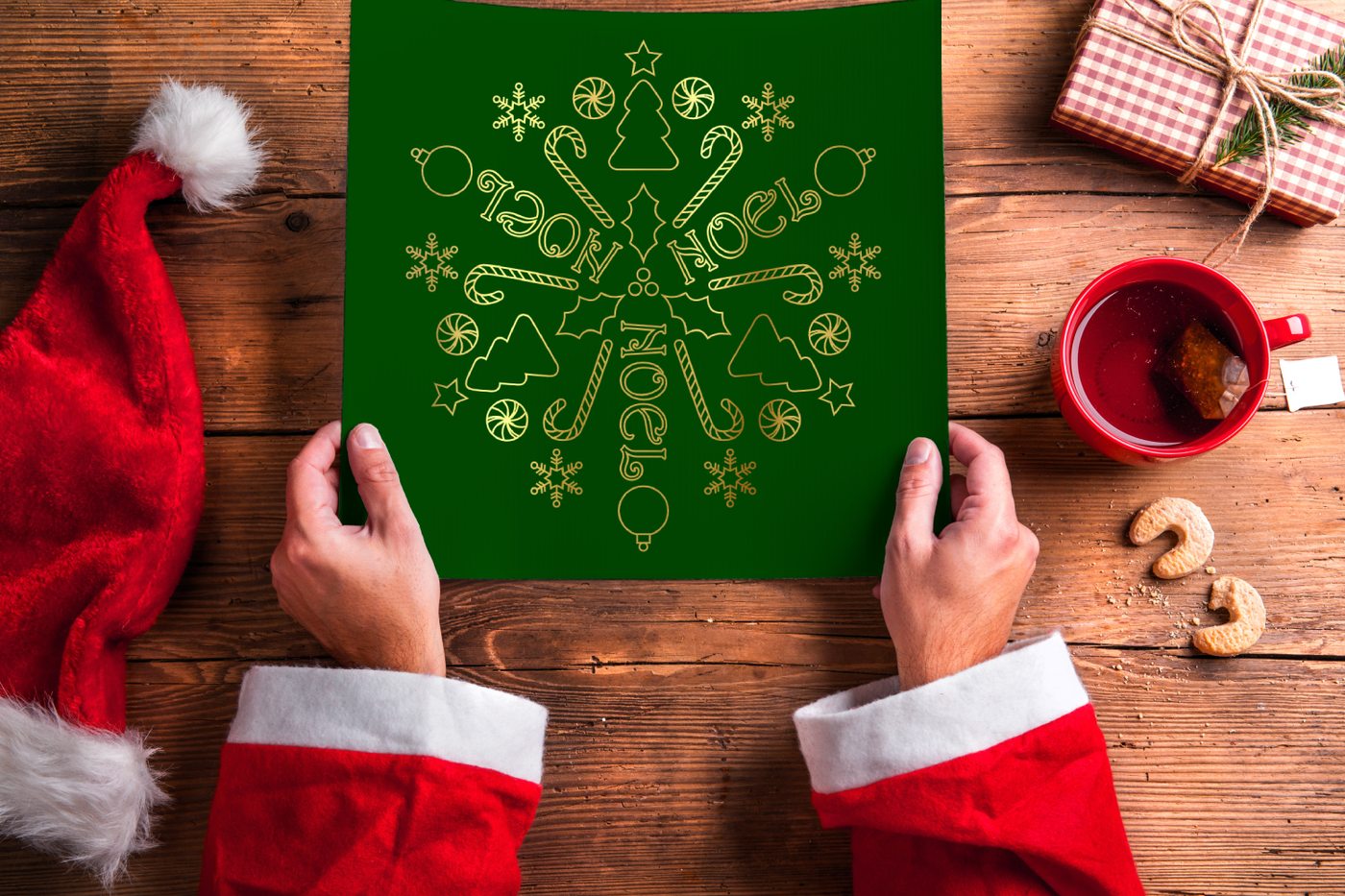 A white person dressed as Santa with only their arms visible holds out a large square piece of green paper. A Santa hat and gift lay to the side. The paper is decorated with the word NOEL and various Christmas icons in gold set on a circle.