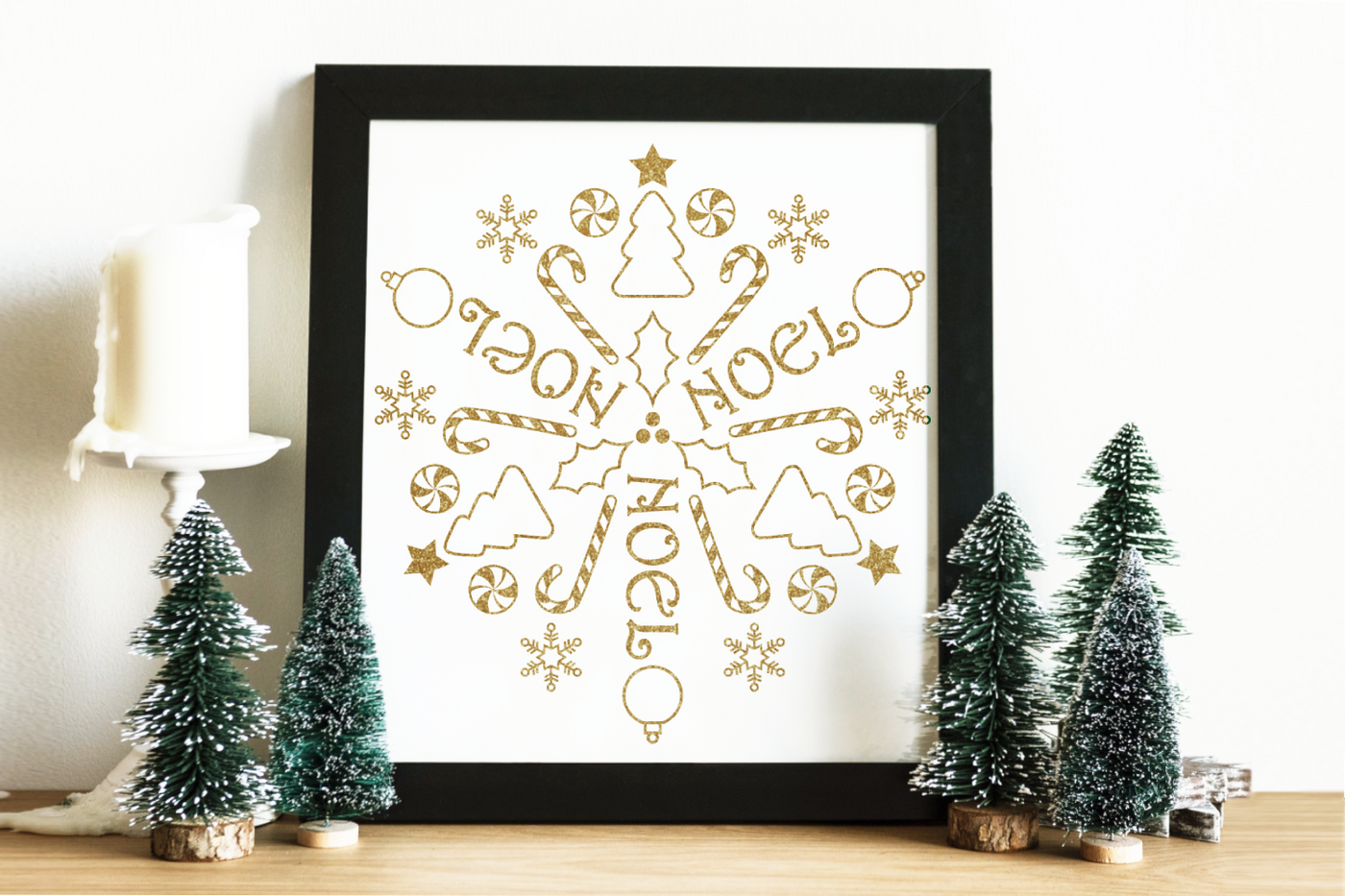 A square framed poster sits on a table with mini Christmas trees to either side. The poster has gold Christmas symbols and the word NOEL in a circular repeat.