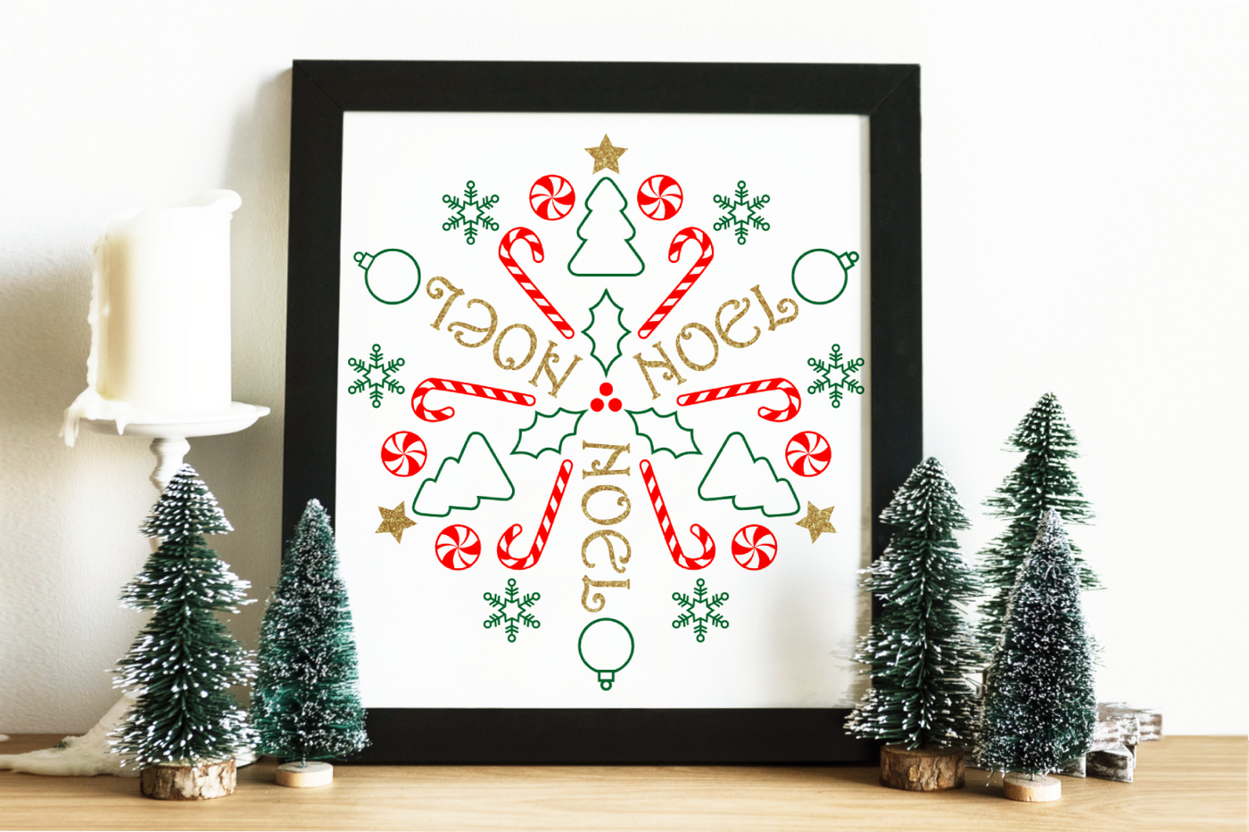 A square framed poster sits on a table with mini Christmas trees to either side. The poster has gold, red, and green Christmas symbols and the word NOEL in a circular repeat.