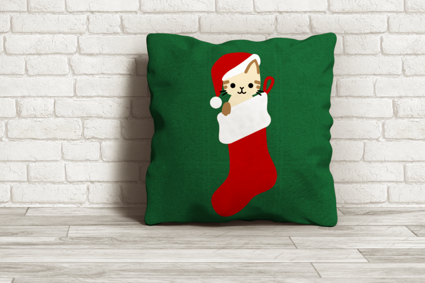 A green throw pillow on the floor in front of a white brick wall. The pillow has a design featuring a kitten in a stocking wearing a Santa hat.
