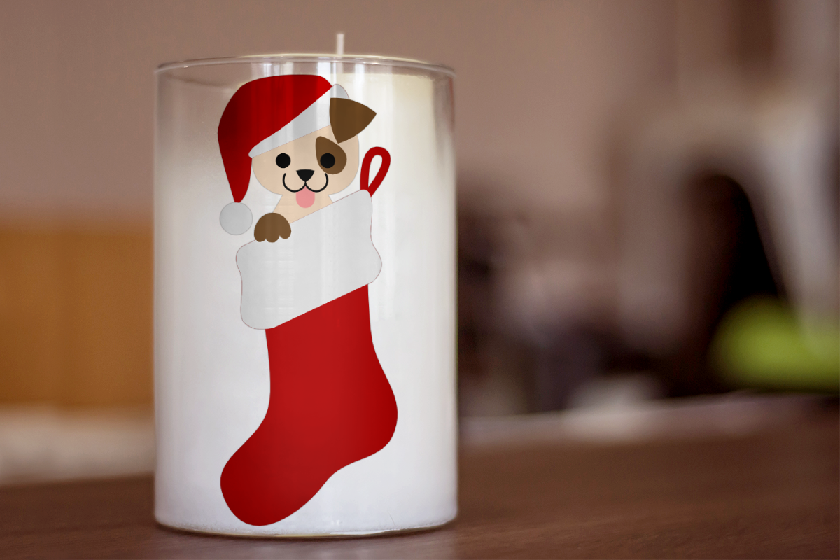 A white candle with the background out of focus. On the candle is a puppy in a stocking wearing a Santa hat.
