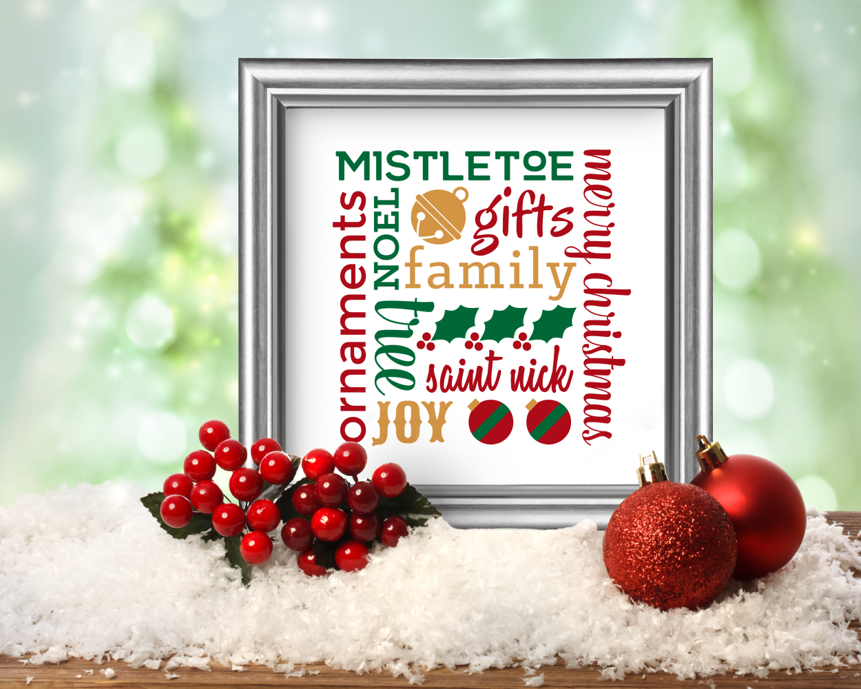 A square framed poster sits on a table with fake snow under it, holly berries, and red Christmas ornaments. The poster has various Christmas words and symbols set on a square in red, green and gold on a white background.