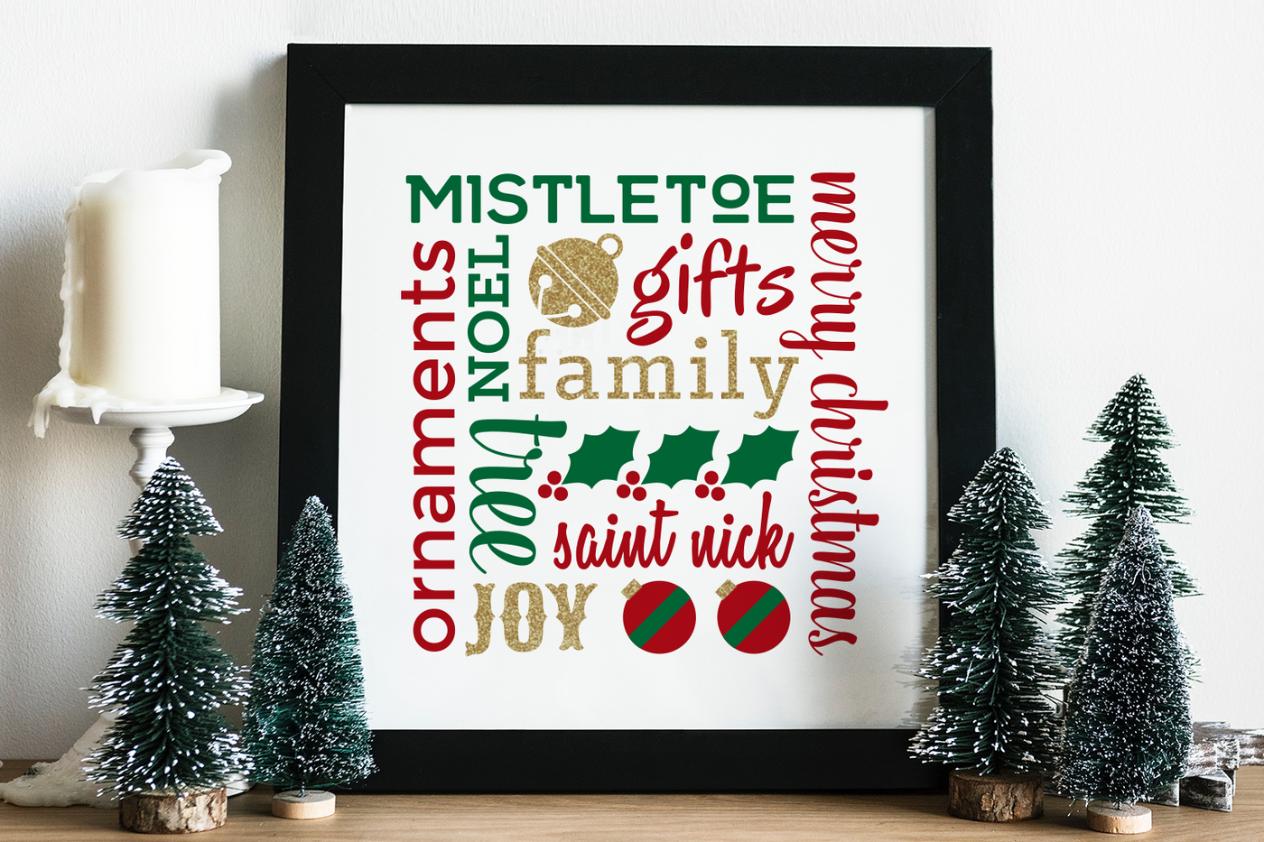 A square framed poster on a table with mini Christmas trees and a candle near it. The poster has various Christmas words and symbols set on a square in red, green and gold on a white background.