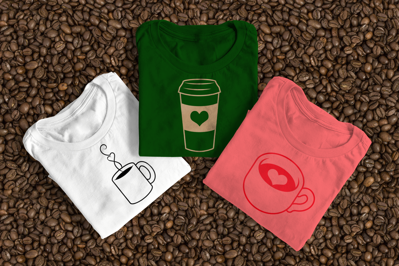 Three folded tees laying on some coffee beans. One has a coffee mug with heart steam, another is a coffee to go cup with a heart on the sleeve, and the last is a latte cup with a heart in the liquid.