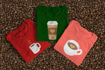 Three folded tees laying on some coffee beans. One has a coffee mug with heart steam, another is a coffee to go cup with a heart on the sleeve, and the last is a latte cup with a heart in the liquid.