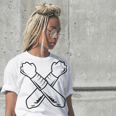 A black women with blonde braids in a high partial bun wears glasses and a white tee. On the tee are the black outlines of two arms crossed with metal wrist cuffs.