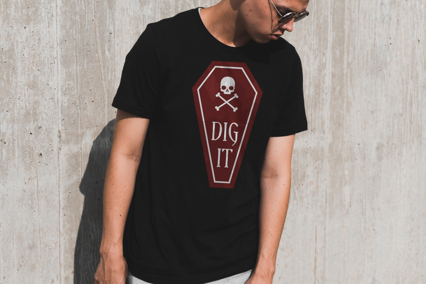 A white young man stands against a cement wall wearing a black tee and sunglasses. The shirt has a dark red coffin with a skull and crossbones and the words "Dig it" in a Gothic font.