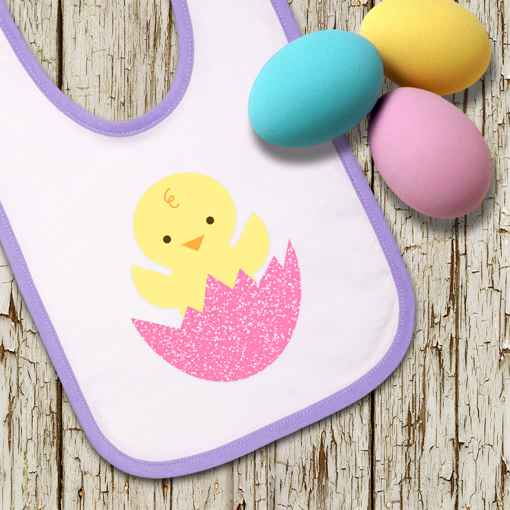 A white bib with lavender trim. Decorating the bib is a baby chick popping out of an egg that is done in pink glitter vinyl. On top of the bib lay pastel colored Easter eggs.