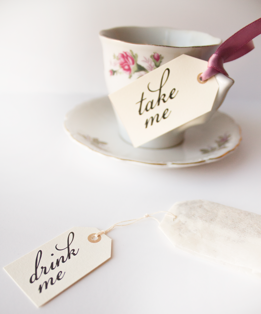 A tea cup with a tag fastened with pink ribbon that says "take me." laying in front of it is a tea bag with a tag that says "drink me."