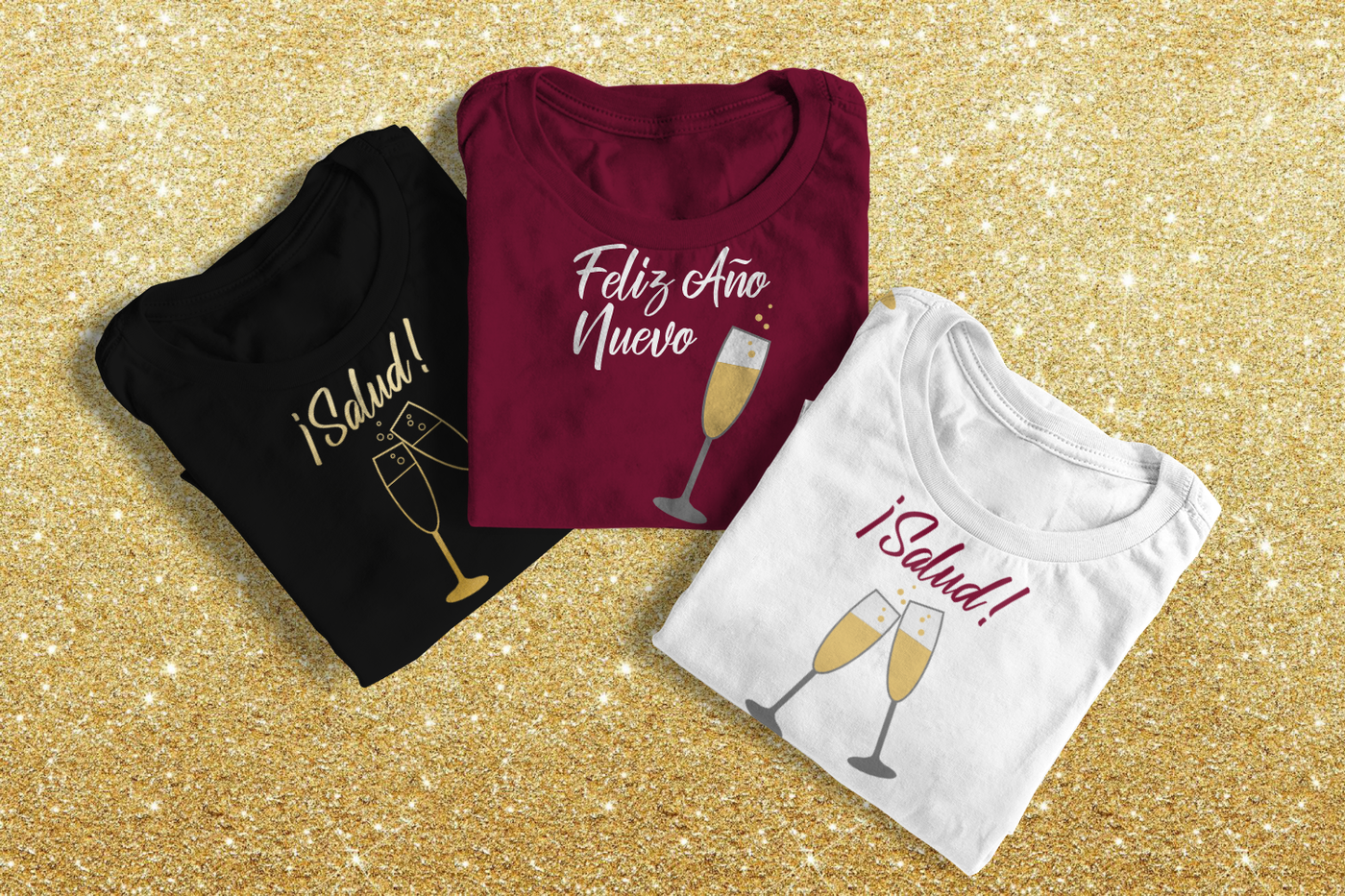 Three folded tees. Two say "Salud!" with toasting champagne glasses. One says "Feliz Año Nuevo" with a single glass of champagne.