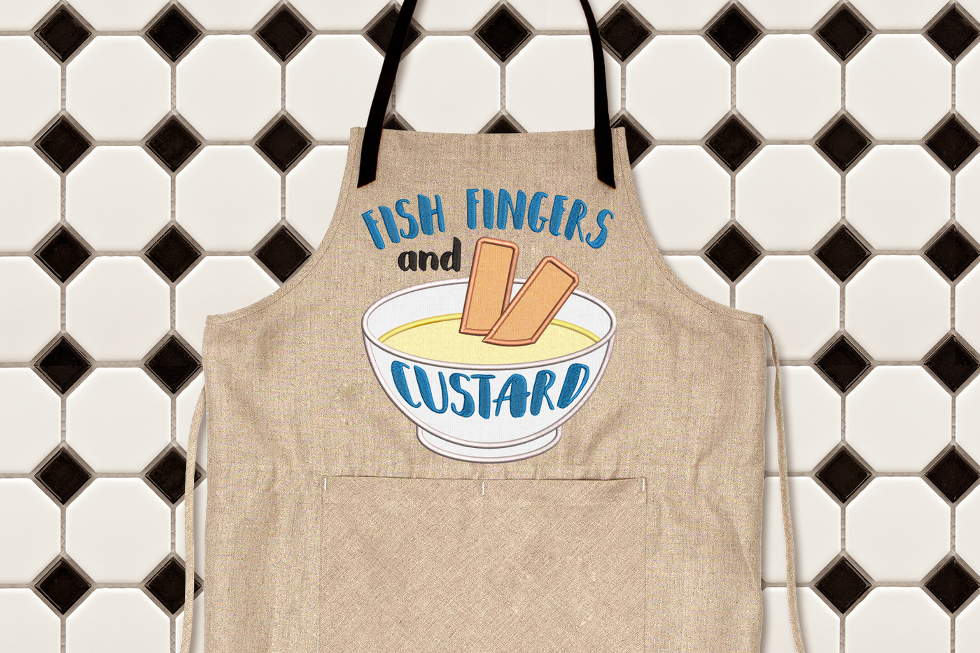 Apron with embroidered words saying "Fish fingers and custard." There is an applique of two fish sticks in a bowl of vanilla pudding.