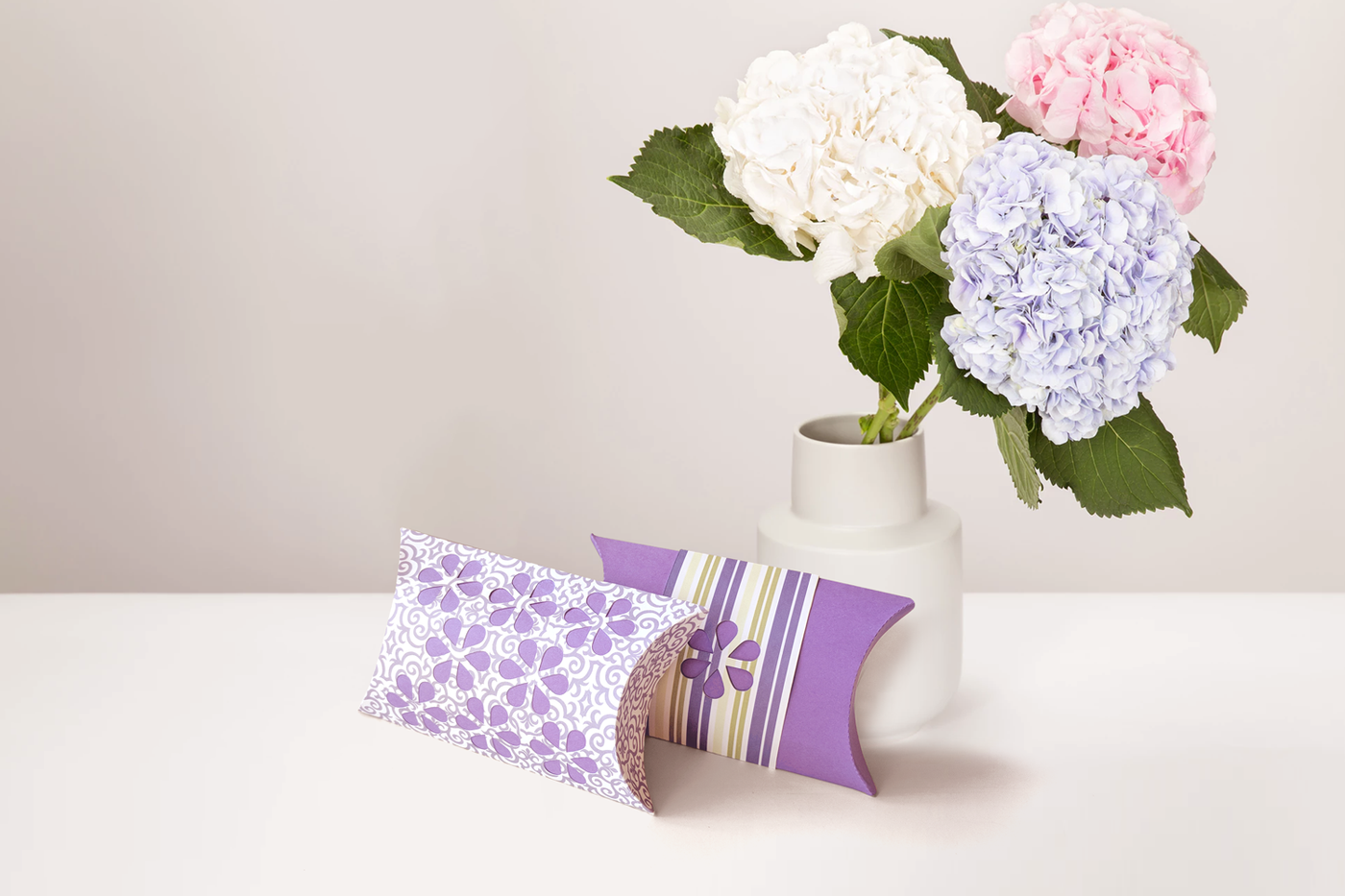 Two pillow boxes in front of a vase of hydrangeas. The front box has a purple and white swirl pattern with flower cutouts to reveal purple paper. The back one is purple with a purple, green, ivory,and white striped band with a flower cutout.
