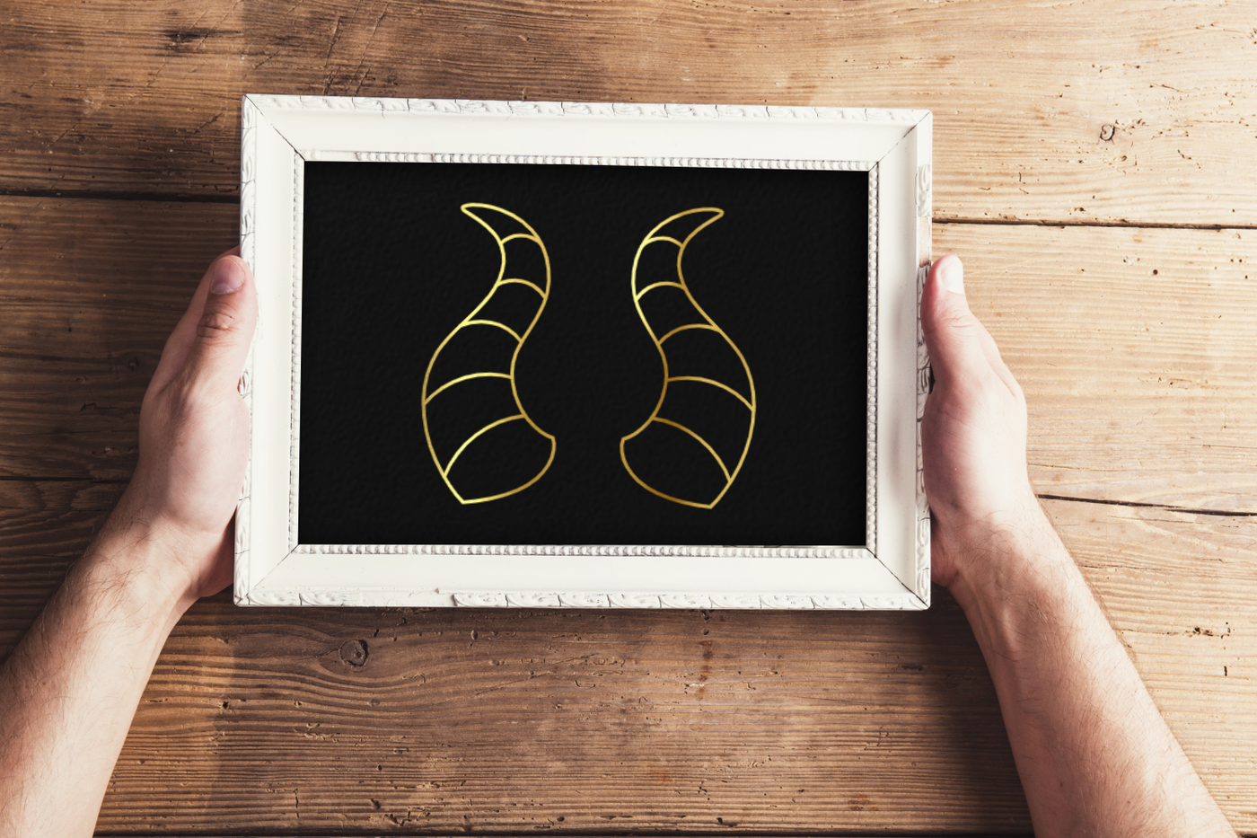 White hands holding a framed image. The image is a gold line drawing of dragon horns on a black background.