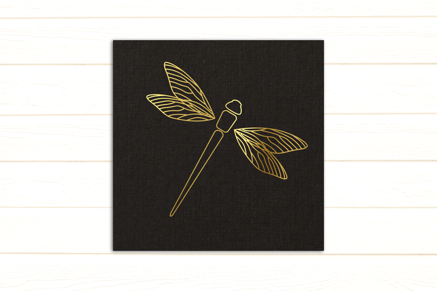 A black piece of paper with the line drawing of a dragonfly with detailed wings. The dragonfly is in gold foil.