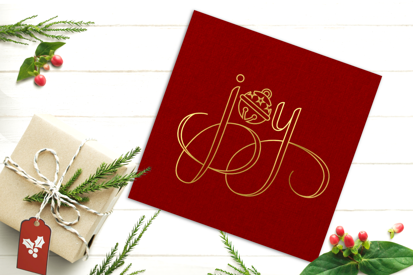 A square red card with the word "joy" in gold. The O is a round bell. Laying near the card are red and green foliage and a brown paper wrapped gift box.