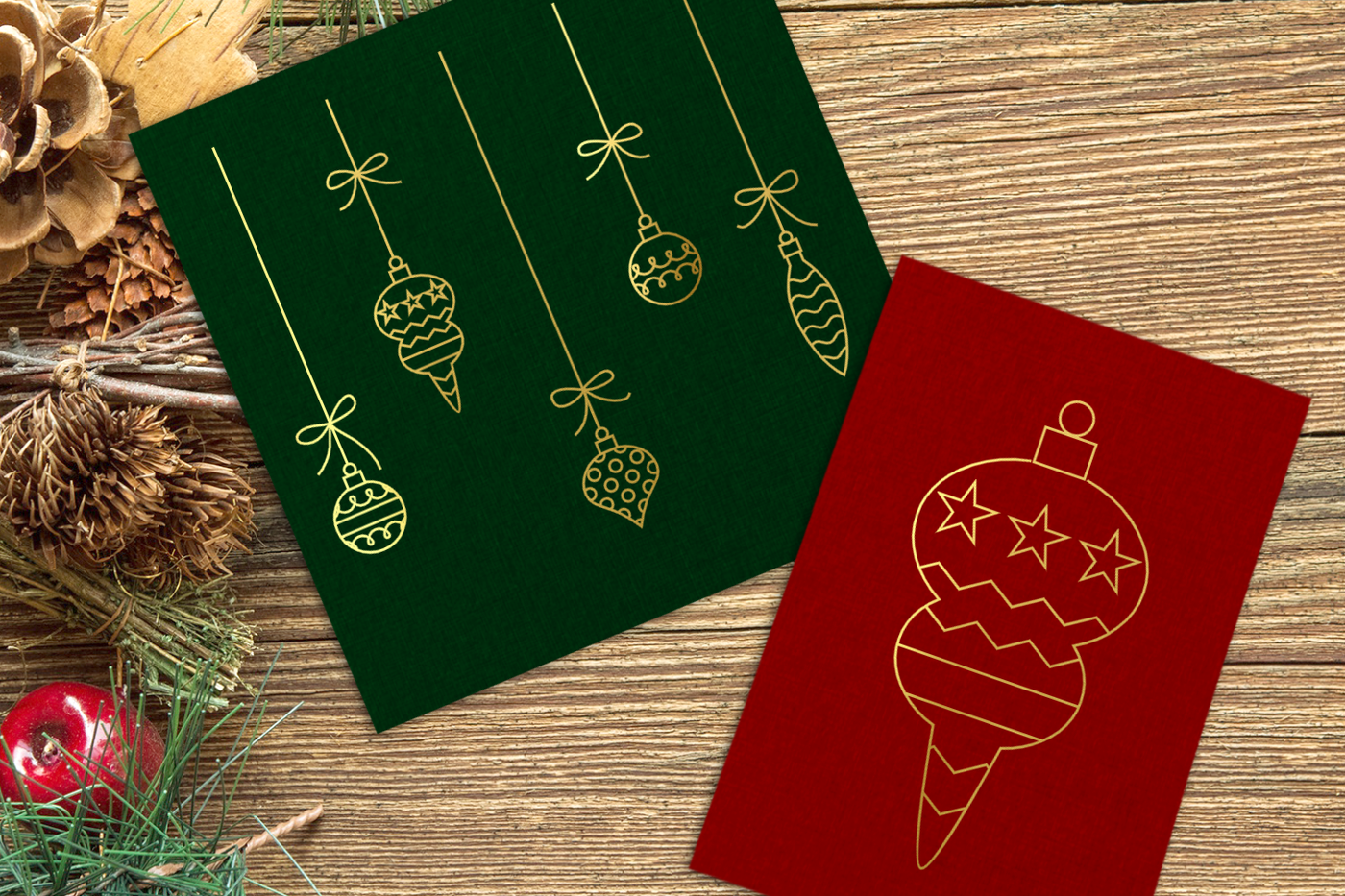 A green and red card sit on a wooden table with holiday greenery nearby. Each has a gold line drawing of holiday ornaments. The green card has multiple ornaments hanging from strings. The red card has a single larger fancy ornament.