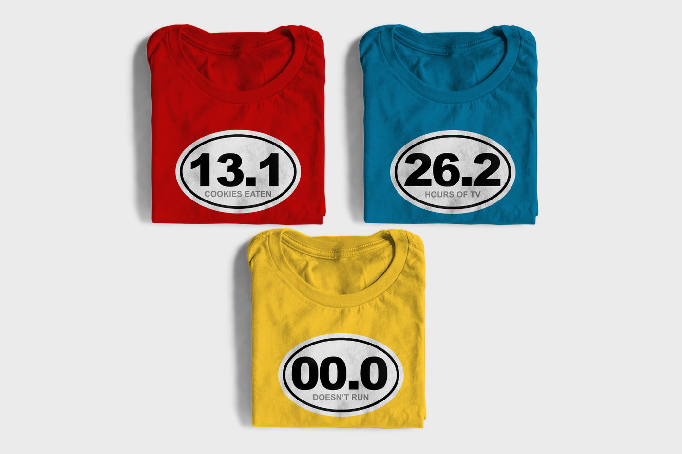 Three folded tees, each with a white oval design. One says "13.1 cookies eaten," one says "26.2 hours of TV," and one says "00.0 doesn't run."