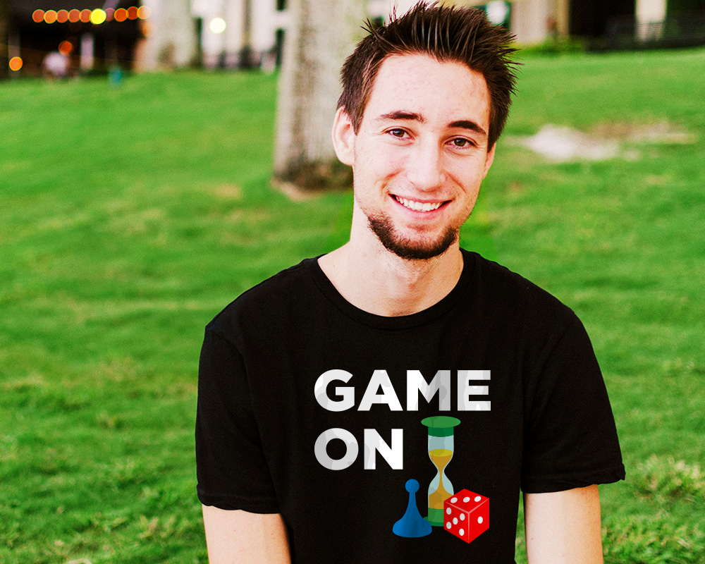 A white man with facial hair on his chin only sits in some grass. He wears a black shirt that says "game on" with a blue game pawn, red 6-sided die, and green sand timer below.