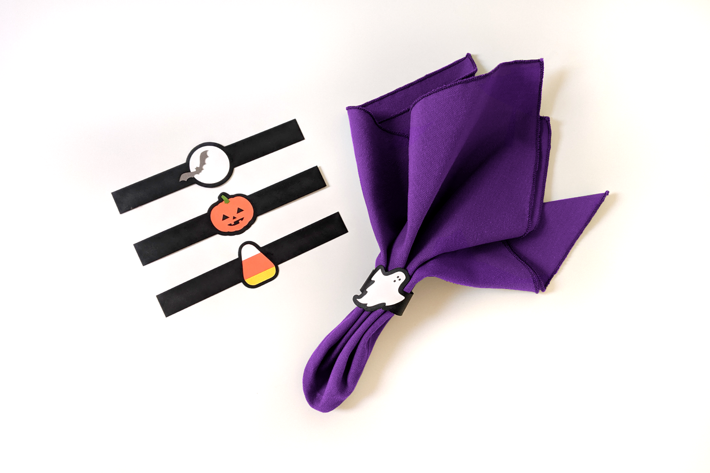 Purple napkin with a paper ghost napkin ring. Laying flat nearby are 3 more paper napkin rings: a bat in front of a full moon, a carved pumpkin, and a candy corn.