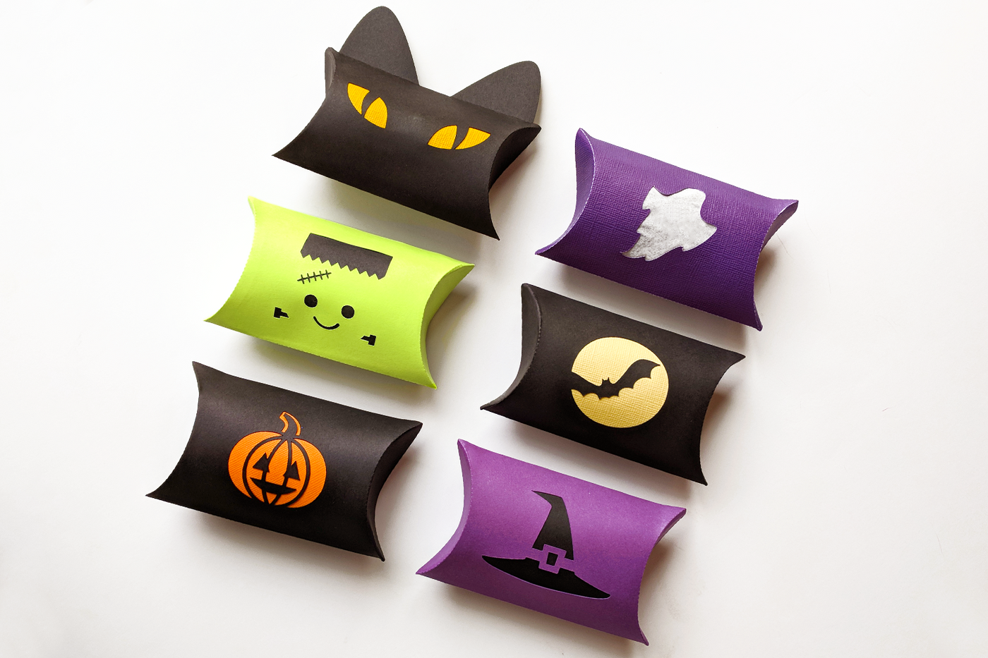 Six pillow boxes in Halloween colors. There is a black cat head, Frankenstein's monster face, a carved pumpkin, ghost, bat in front of a full moon, and witch hat.