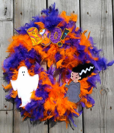 A purple and orange feather wreath with 3 different Halloween applique designs added - a ghost, Bride of Frankenstein, and the word "BOO!"n