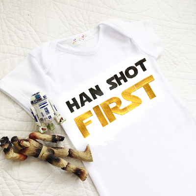 Baby onesie embroidered with "Han shot first" in black and gold. Star Wars action figures lay nearby.