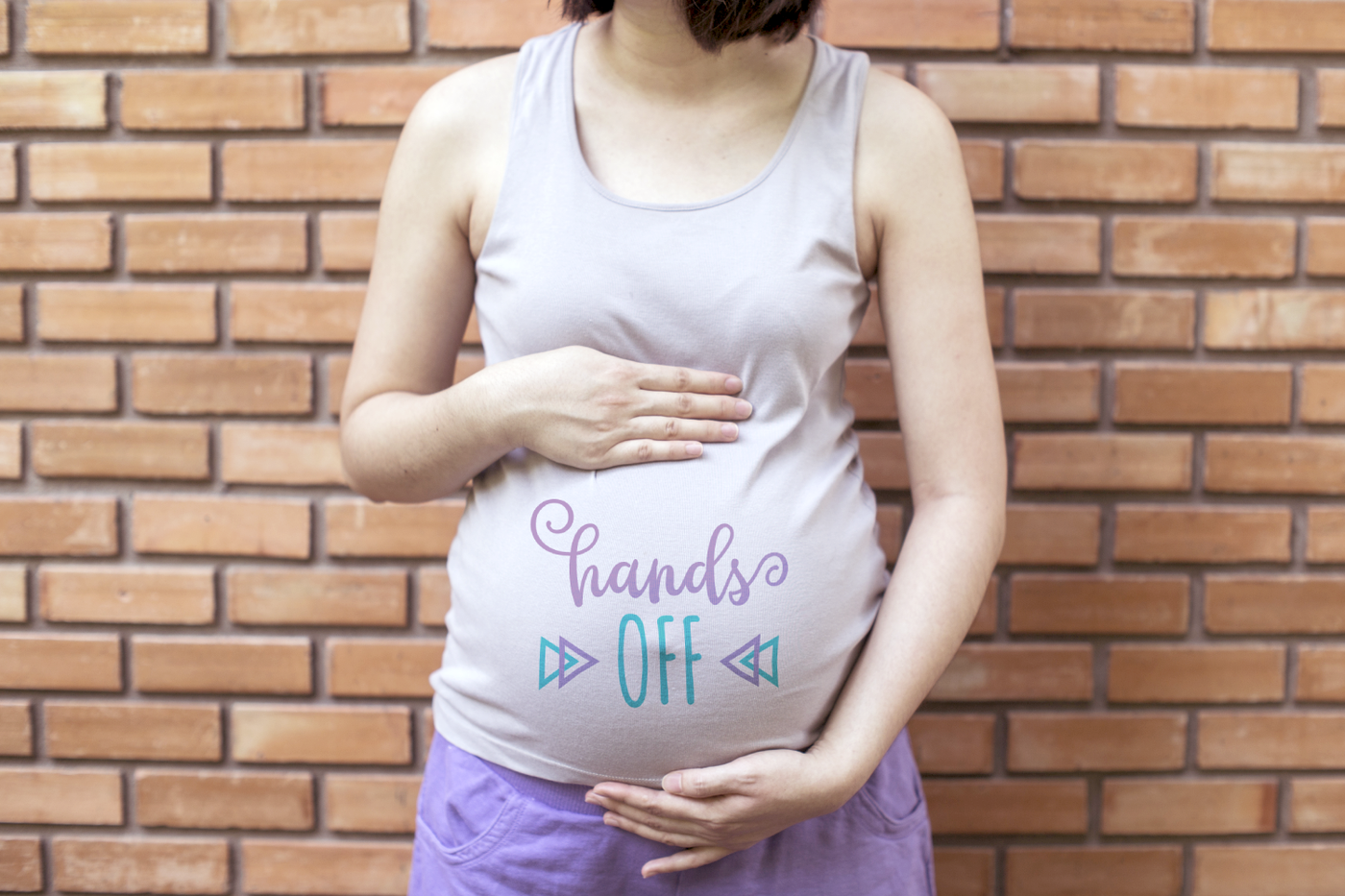 Pregnant white person with their hands above and below their belly. On their baby bump, it says "hands OFF" with decorative triangles on either side.