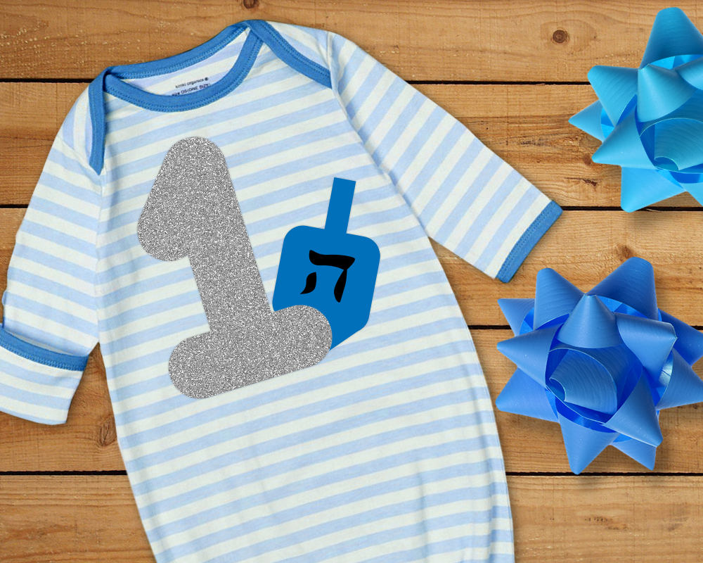 A blue and white striped baby nightgown. On it is a large silver 1 with a blue dreidel behind it.