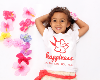 Black little girl lays among tropical flowers. Her shirt has a hibiscus flower design with the words "happiness is where you are."