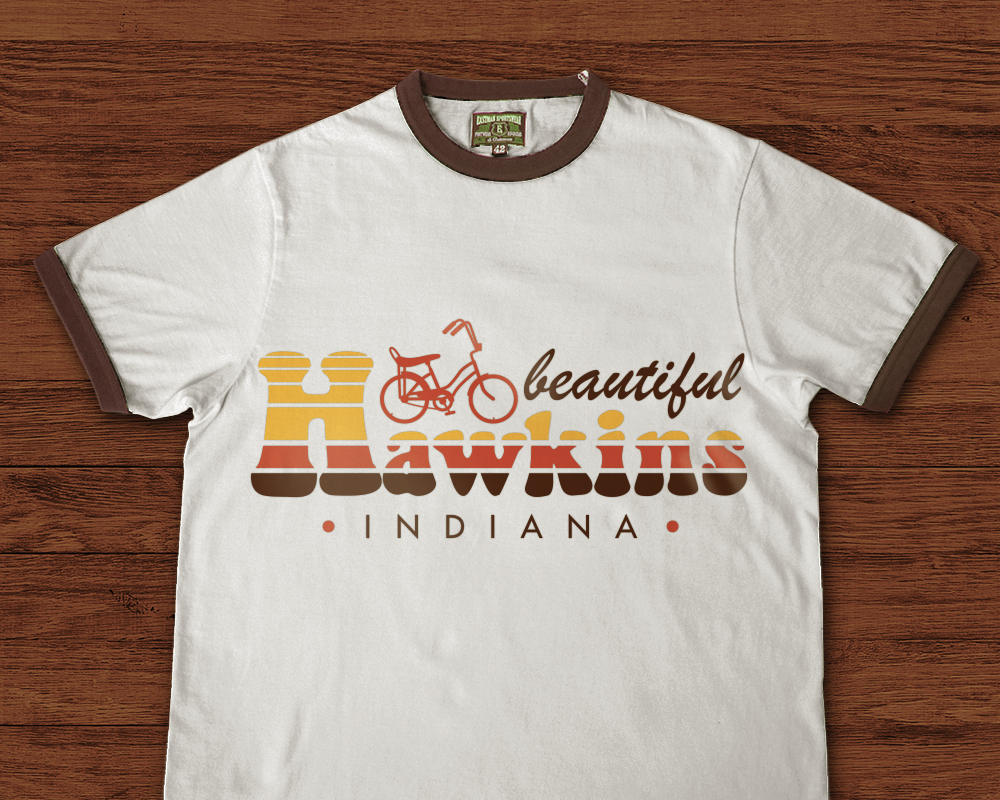 A white ringer tee with brown trim on a retro wood paneled background. The shirt says "beautiful Hawkins Indiana" in retro sunset stripes in yellow, orange, and brown. also has a retro style bicycle at the top.