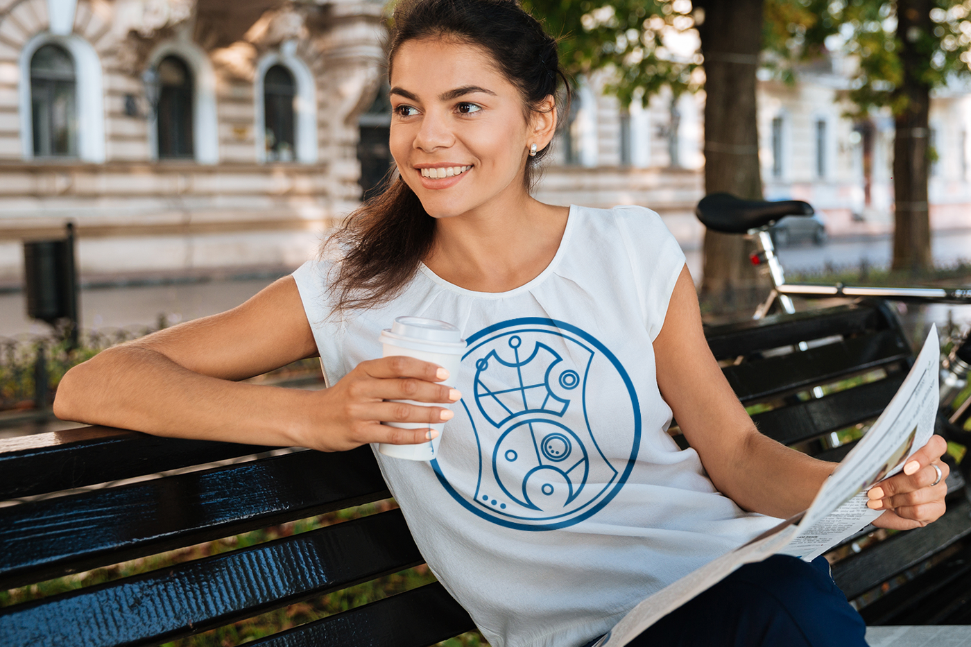 Woman of color in a white shirt sitting on a bench. He shirt has a circular alien language design in blue.