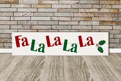 A horizontal sign lays against a brick wall. The sign has holly on it and says "Fa La La La La."
