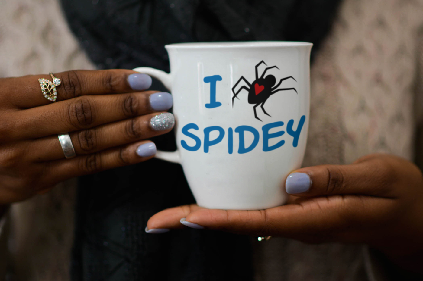 A black woman with lavender nail polish holds a white mug. On the mug, it says "I heart spidey" in comic book style letters. The heart is on the back of a crawling spider design.